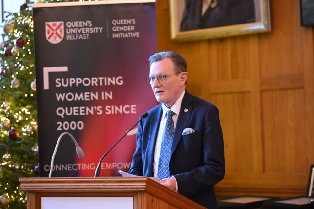 man wearing suit and glasses speaking from a podium. A pop-up stand in the background reads 'Queen's University Belfast / Queen's Gender Initiative / Supporting Women in Queen's since 2000'.