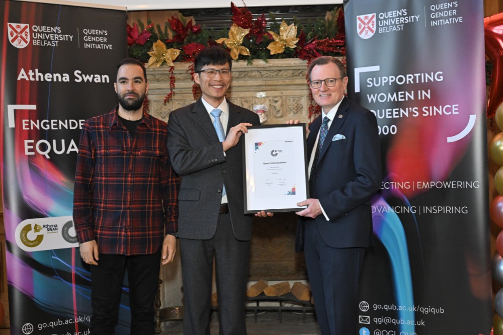 two smiling besuited men posing with a colleague and holding a framed painting in an old event hall with large fireplace in the background and pop-up stands and balloon pillars on either side