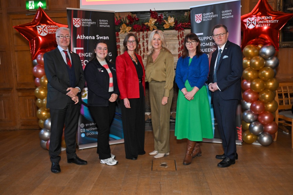 group of two men and four women posing for a photo in an old wood-paneled event hall with pop-up stands and presentation screen in the background