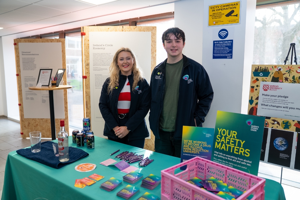 Two student union staff members stand behind a campus safety information table displaying leaflets and materials, with exhibition boards in the background.