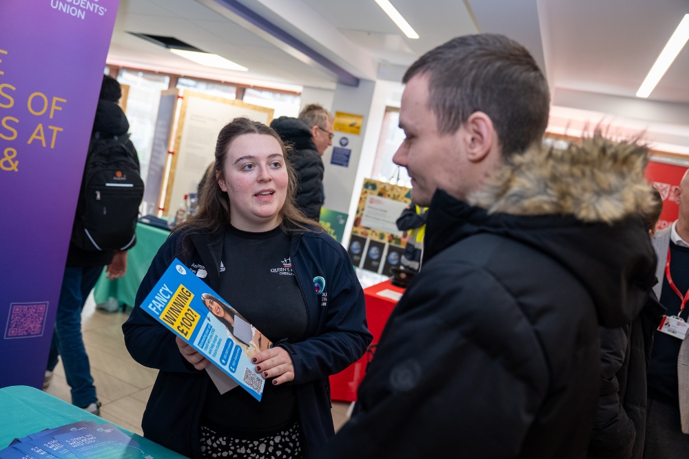 Student Union President Amy Smith  speaks with an attendee at an indoor campus event while holding an information leaflet.