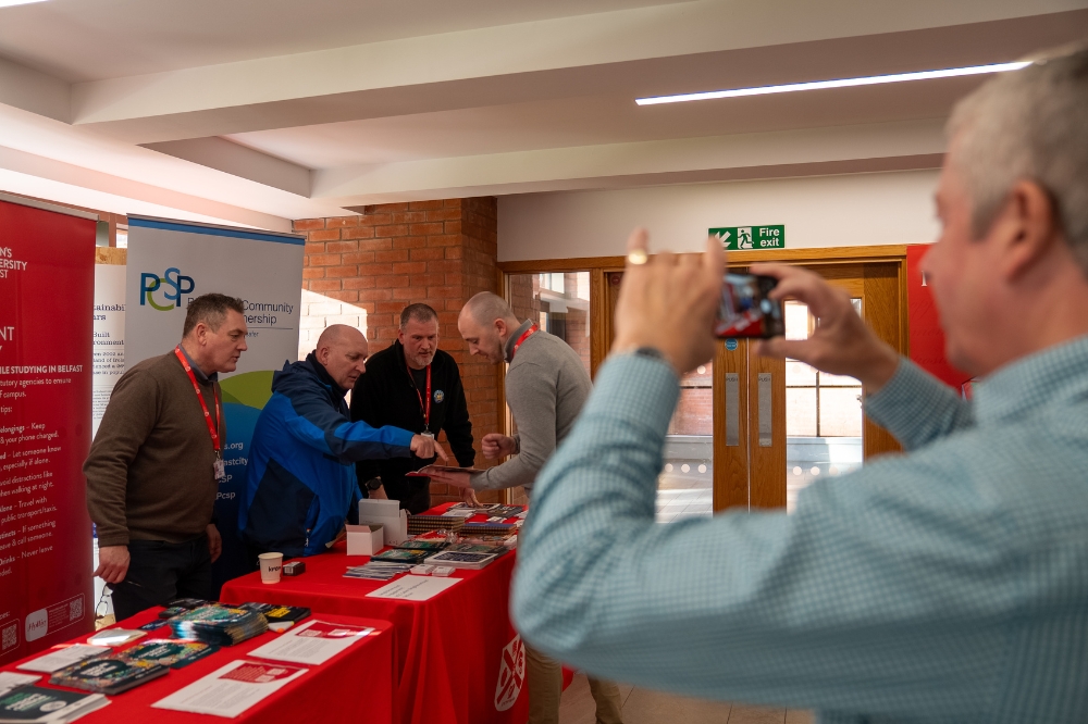 People gathered around a campus safety information stall as one person takes a photo on a smartphone; staff stand behind a table with leaflets and materials, with Queen’s University Belfast and community safety banners in the background.