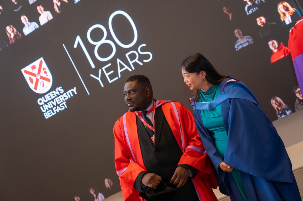 two academic staff wearing graduation robes standing in front of a big screen showing the words 'Queen's University Belfast, 180 Years'