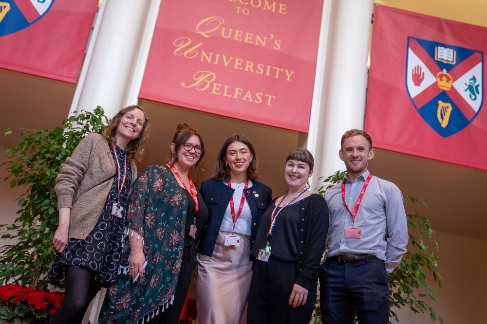 group of four women and a man smiling to camera in an indoor space with a banner above them, flanked by university crests, that reads 'Welcome to Queen's University Belfast'