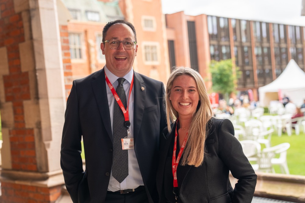 man and woman in business dress wearing red lanyards smiling to camera with lawns and a building in the background