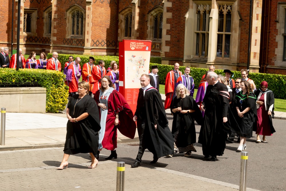 an academic graduation procession turning a corner outside a redbrick building