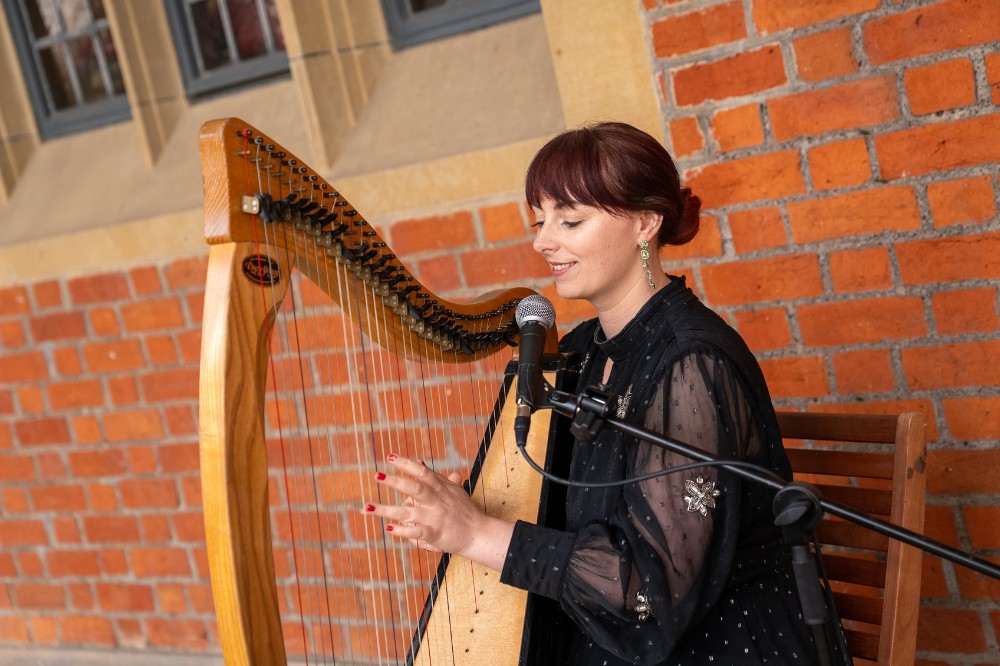 young woman in a black jacket playing a harp with a redbrick wall behind her