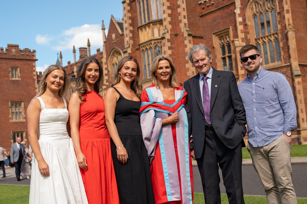 mature lady in graduation robe surrounded by family and posing for a photo with a redbrick building in the background