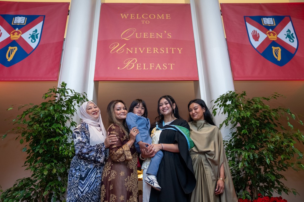 four women, one wearing graduation robe holding a child, standing in the foyer of a large hall with a crest-flanked banner above them reading 'Welcome to Queen's University Belfast'