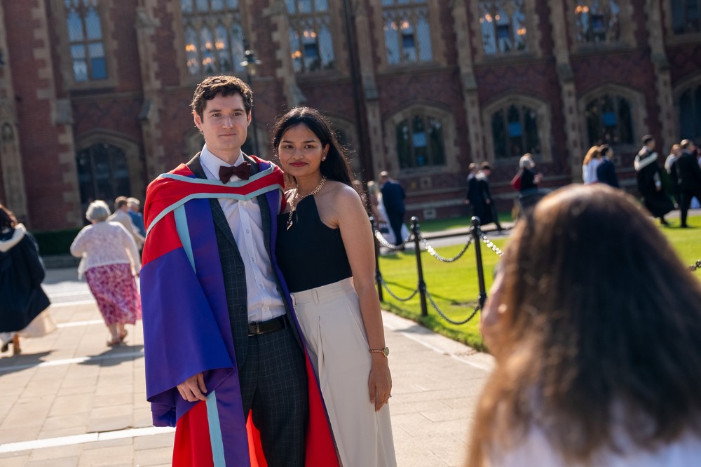 young man in graduation robe standing with his partner and smiling as someone takes their photo on a sunny day in front of a redbrick building
