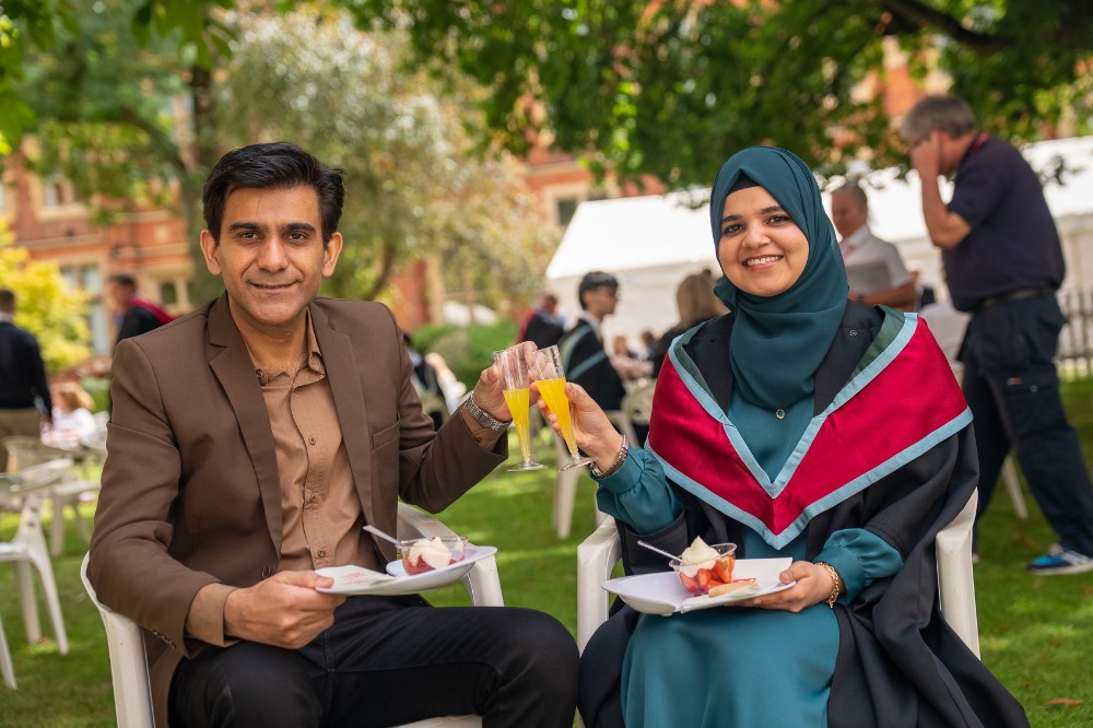 seated man and woman of Eastern origin clinking glasses of orange and enjoying strawberries and cream on a lawn area with trees - the women is wearing a graduation robe