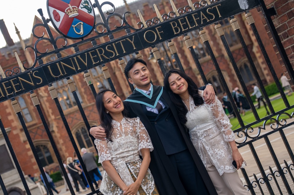 young international man in graduation robe flanked by two young women posing for photos outside large gates reading 'Queen's University Belfast'