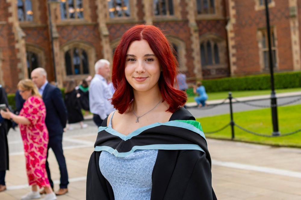 young woman in light blue dress and graduation robe smiling to camera with a redbrick building in the background