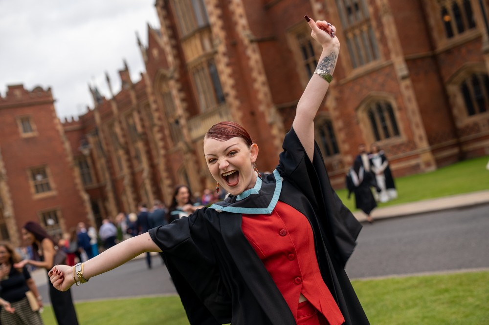 young woman in red dress and graduation robe striking a celebratory pose with a redbrick building in the background