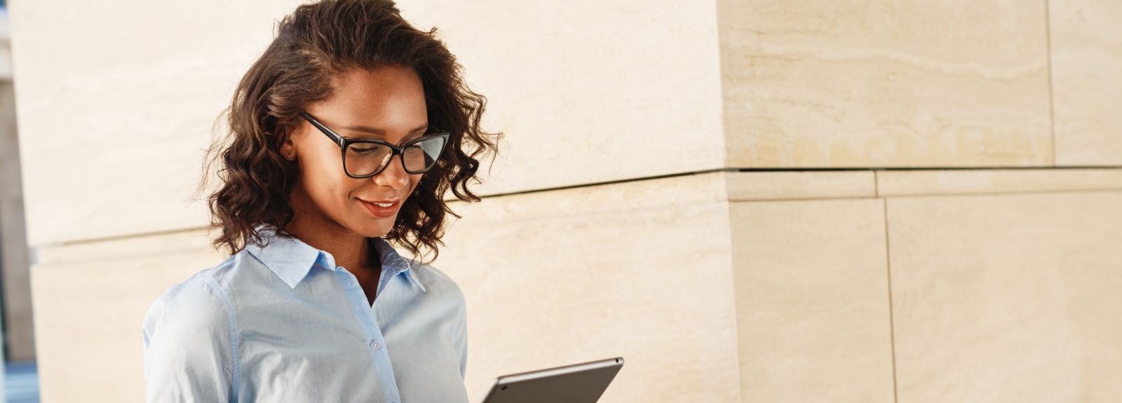 A woman wearing glasses and a light blue shirt stands outdoors reading a tablet, smiling slightly.