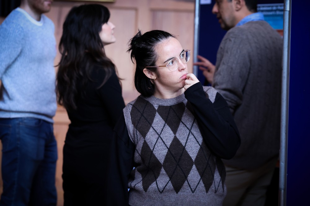 Woman considering a poster at an indoor research poster exhibition. Two other people are pictured chatting in the background.