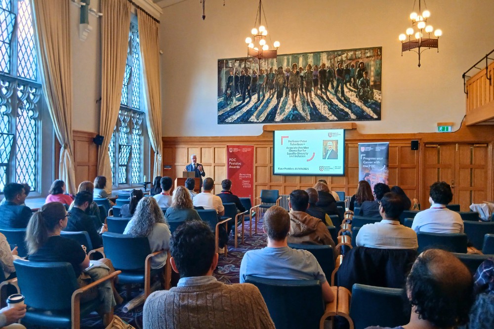 view from the back of a comfortable-looking, wood-paneled meeting room showing group of seated people listening to a speaker at a podium. A large painting hangs on the back wall.