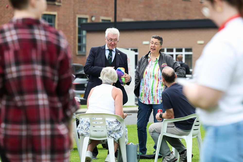 two people standing and chatting to another person seated at an outdoor event on a lawn