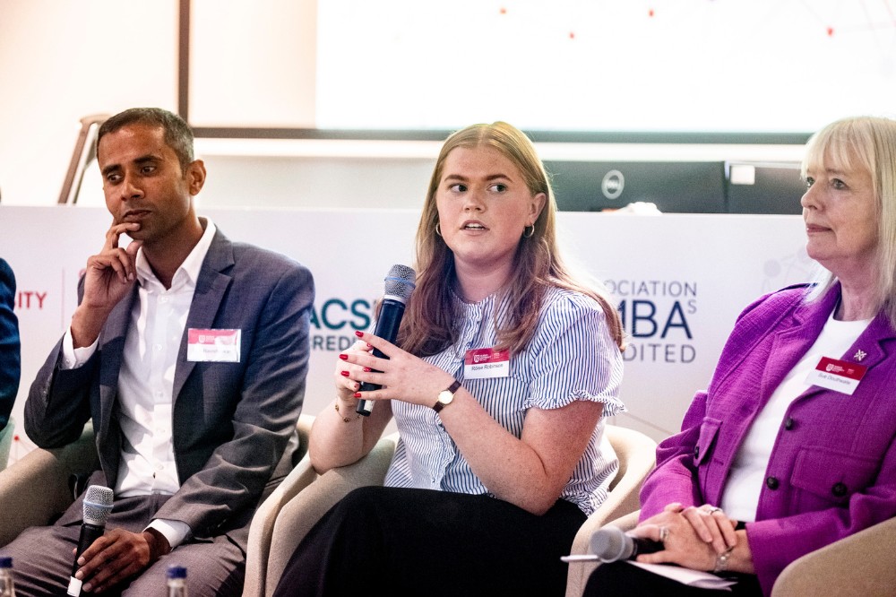 young female panelist speaking into a microphone with two other male and female panelists shown listening on either side