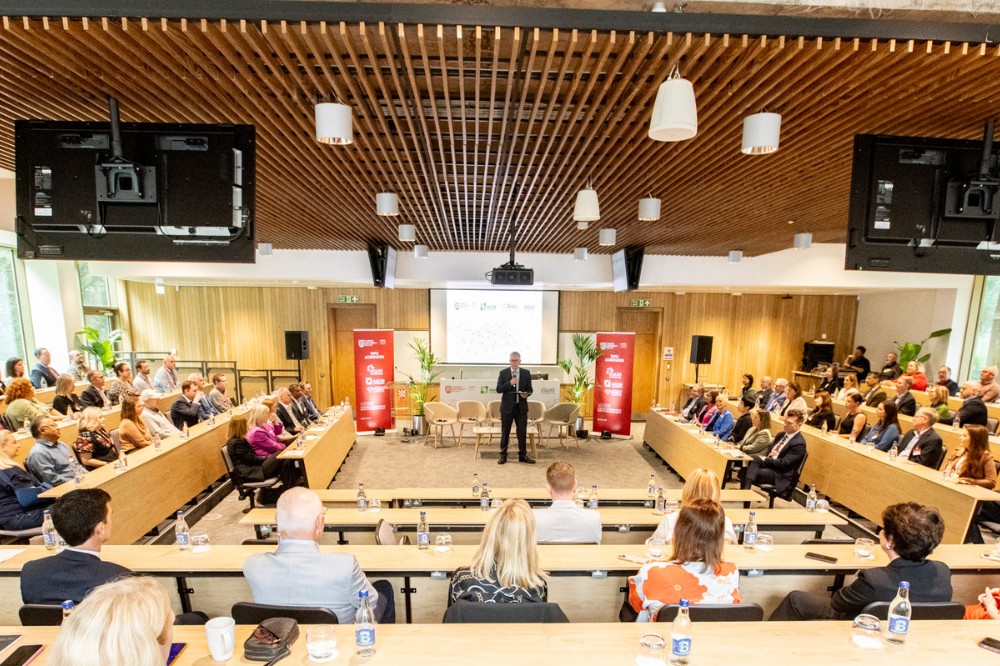 bright, modern lecture hall viewed from the back with presenter shown speaking at the front and seated guests listening in