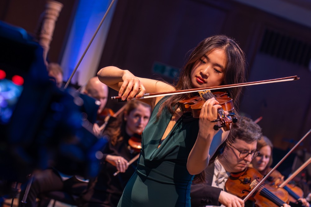 young woman violin soloist standing playing with an orchestra, some of whom are pictured behind her