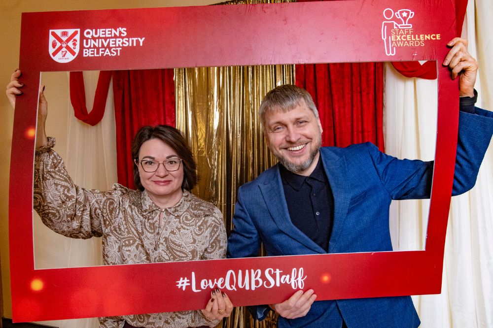 smiling man in blue jacket and black shirt and woman wearing Paisley-patterned blouse posing with an Instagram photo frame cut-out