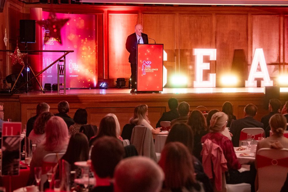 a man speaking to an audience from a podium on a raised stage at an indoor event