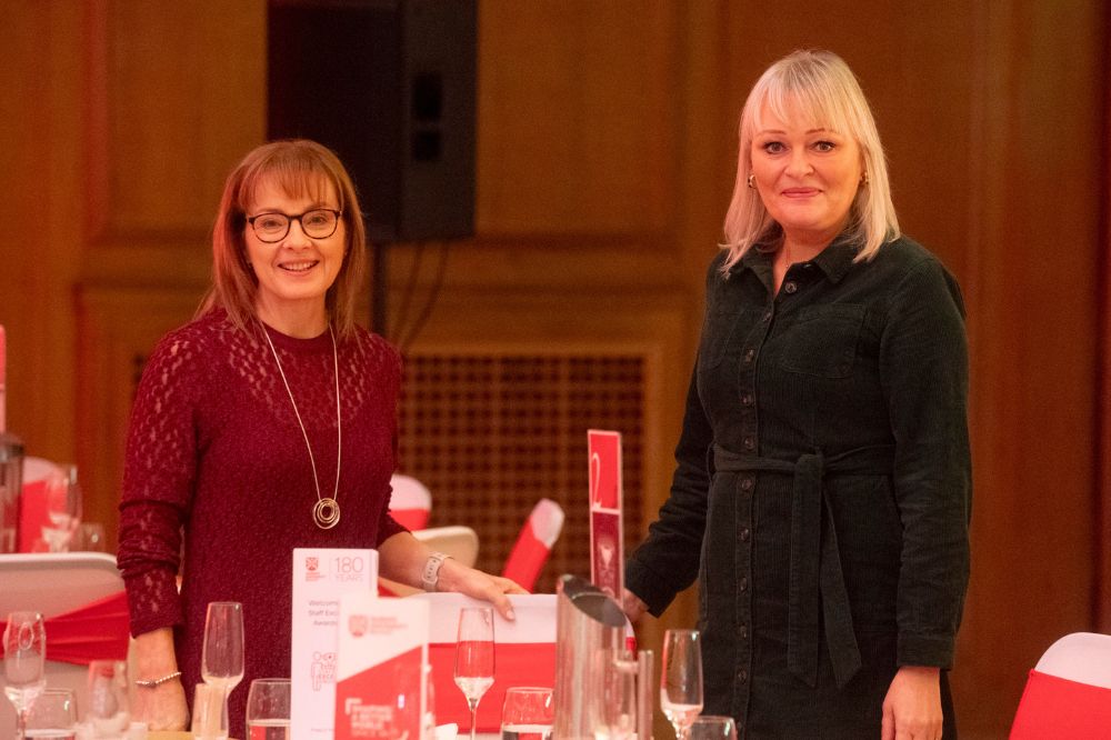 two smiling women posing for the camera while standing at a set dining table at an indoor event