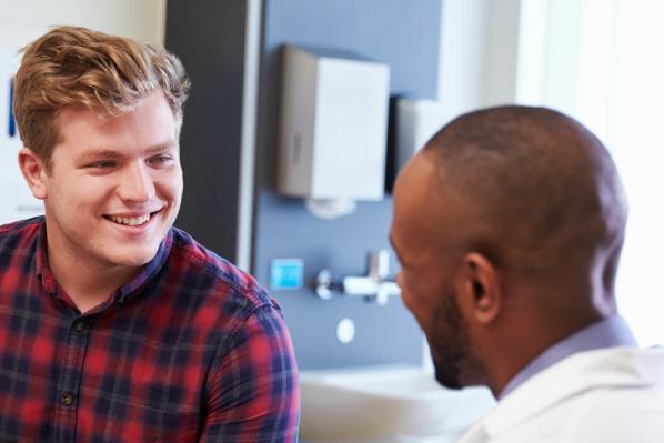 A young man wearing a red check shirt smiles while talking with a healthcare professional in a bright consultation room.