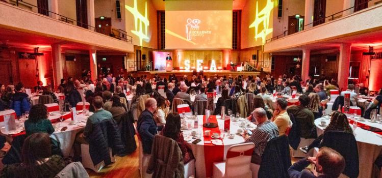 event in a large colourful hall with groups of people sitting at dining tables watching someone speaking from a podium on a stage in the background