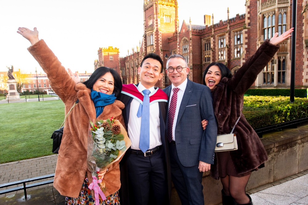 young international man in graduation robe celebrating with his family in front of an old redbrick building