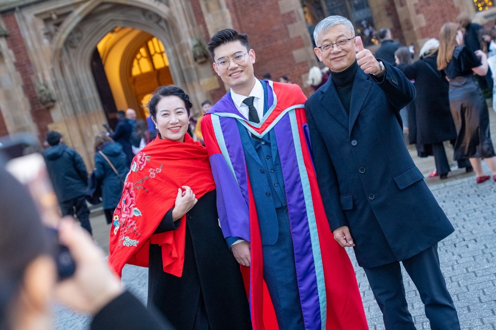young Asian man in red and blue graduation robe celebrating his achievement with his parents while someone takes a picture in the foreground